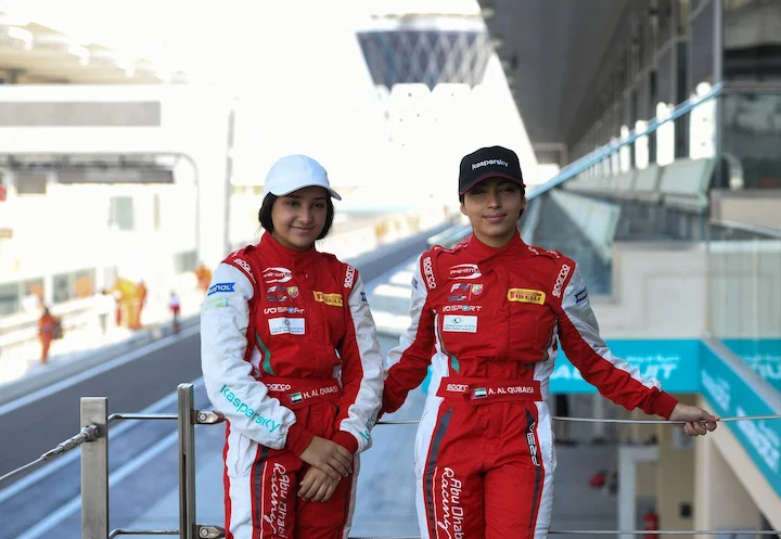Amna and Hamda Al Qubaisi stand side by side at a racetrack in full racing gear.