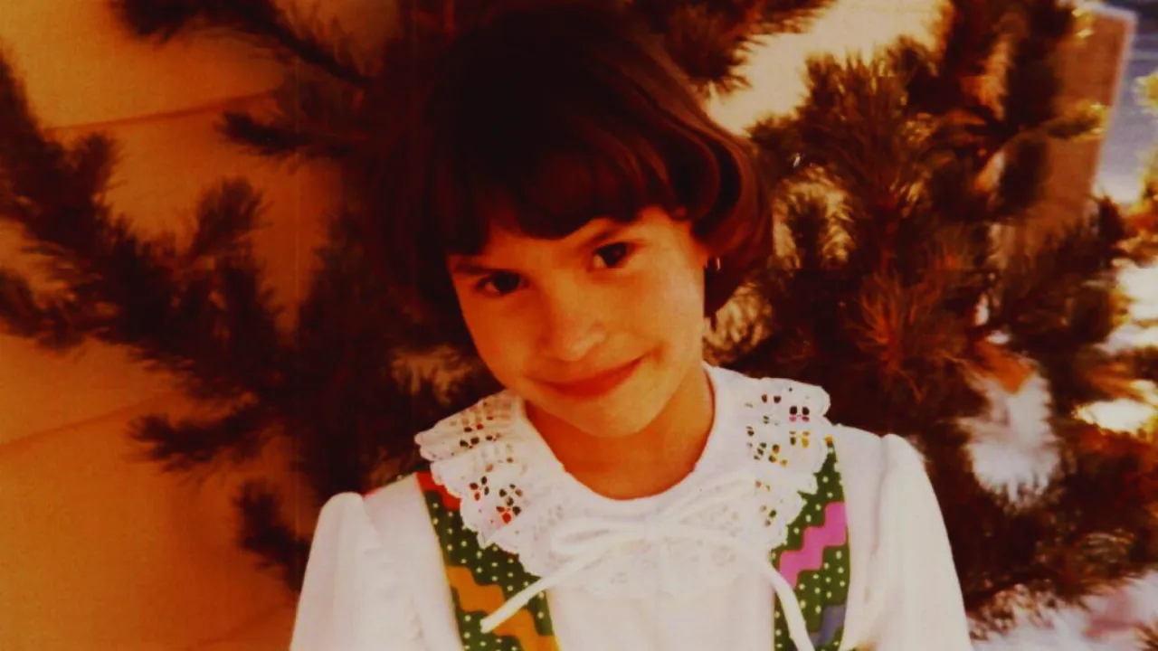 Childhood photo of Jonelle Matthews smiling outdoors in a white blouse with green and pink trim.
