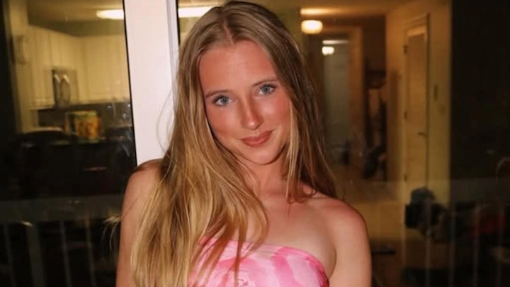 Anna Phelan posing in a pink top with long blonde hair inside a cozy indoor setting.