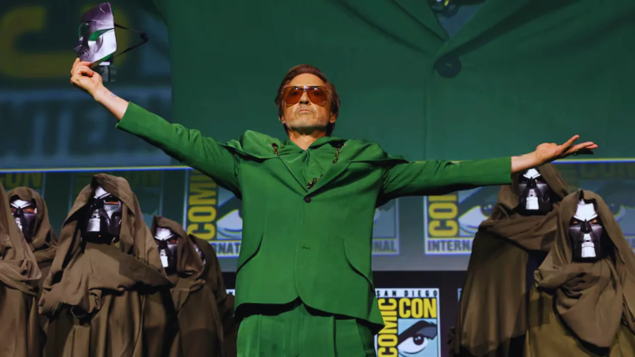 Robert Downey Jr. in green suit raises arms at Comic-Con surrounded by Doctor Doom cosplayers.