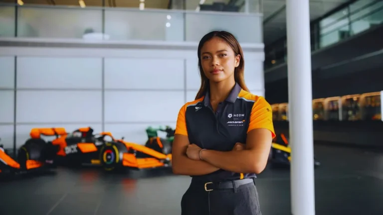 Bianca Bustamante stands in McLaren gear with F1 cars behind her at a racing facility.