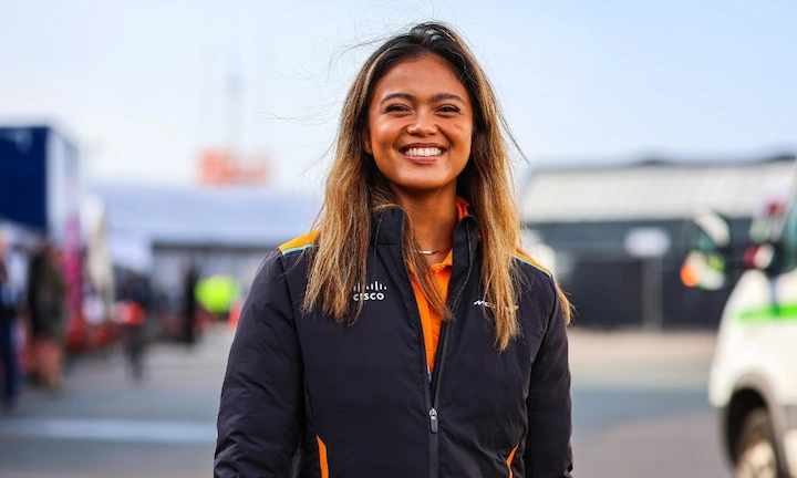 Bianca Bustamante smiles in a McLaren jacket at a racetrack with pit lane in background.