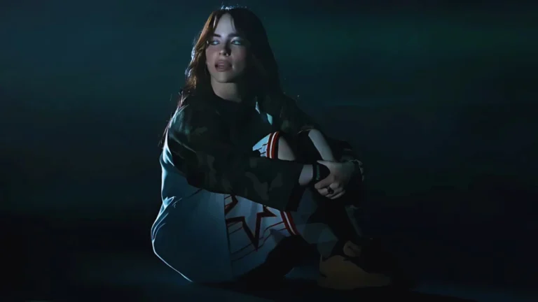 Billie Eilish sitting in a moody spotlight, wearing a camo jacket and patterned shorts, looking reflective.