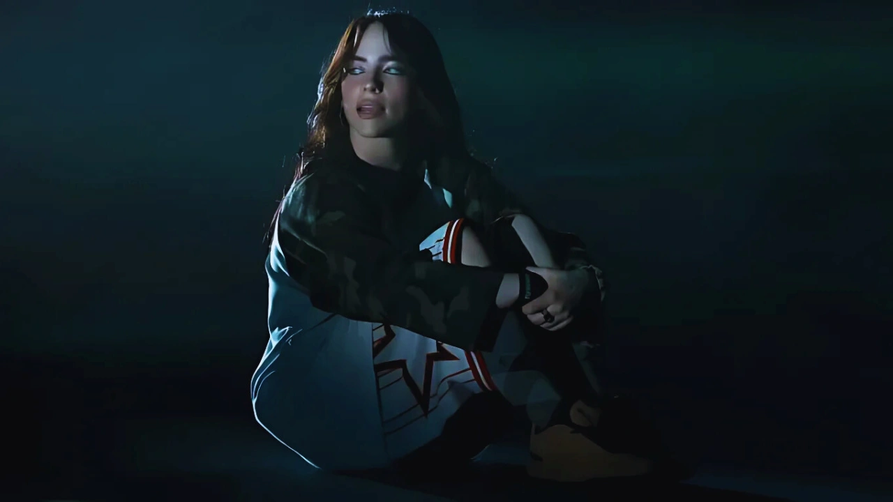 Billie Eilish sitting in a moody spotlight, wearing a camo jacket and patterned shorts, looking reflective.