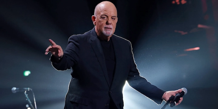 Billy Joel performing on stage in a black suit with microphone in hand.