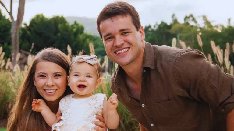 Bindi Irwin smiling with husband Chandler Powell and daughter Grace in an outdoor family photo.