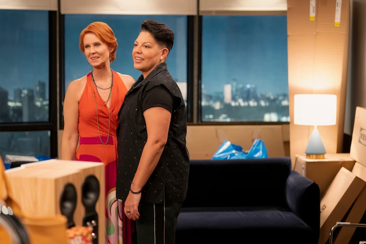 Cynthia Nixon and Sara Ramirez standing together during a scene from And Just Like That, surrounded by moving boxes in a modern apartment.