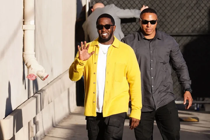 Sean “Diddy” Combs wearing a yellow jacket, waving, accompanied by a bodyguard in a public setting.
