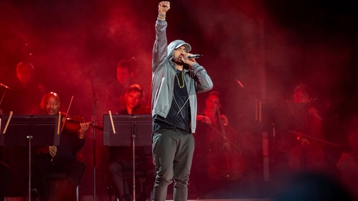 Eminem on stage raising his fist during a dramatic performance with a red-lit orchestra in the background.