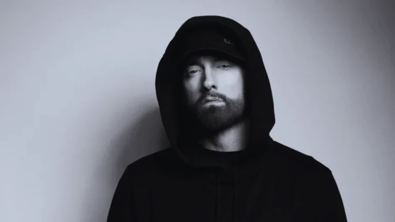 Eminem wearing a black hoodie and hat, staring directly at the camera with a serious expression.