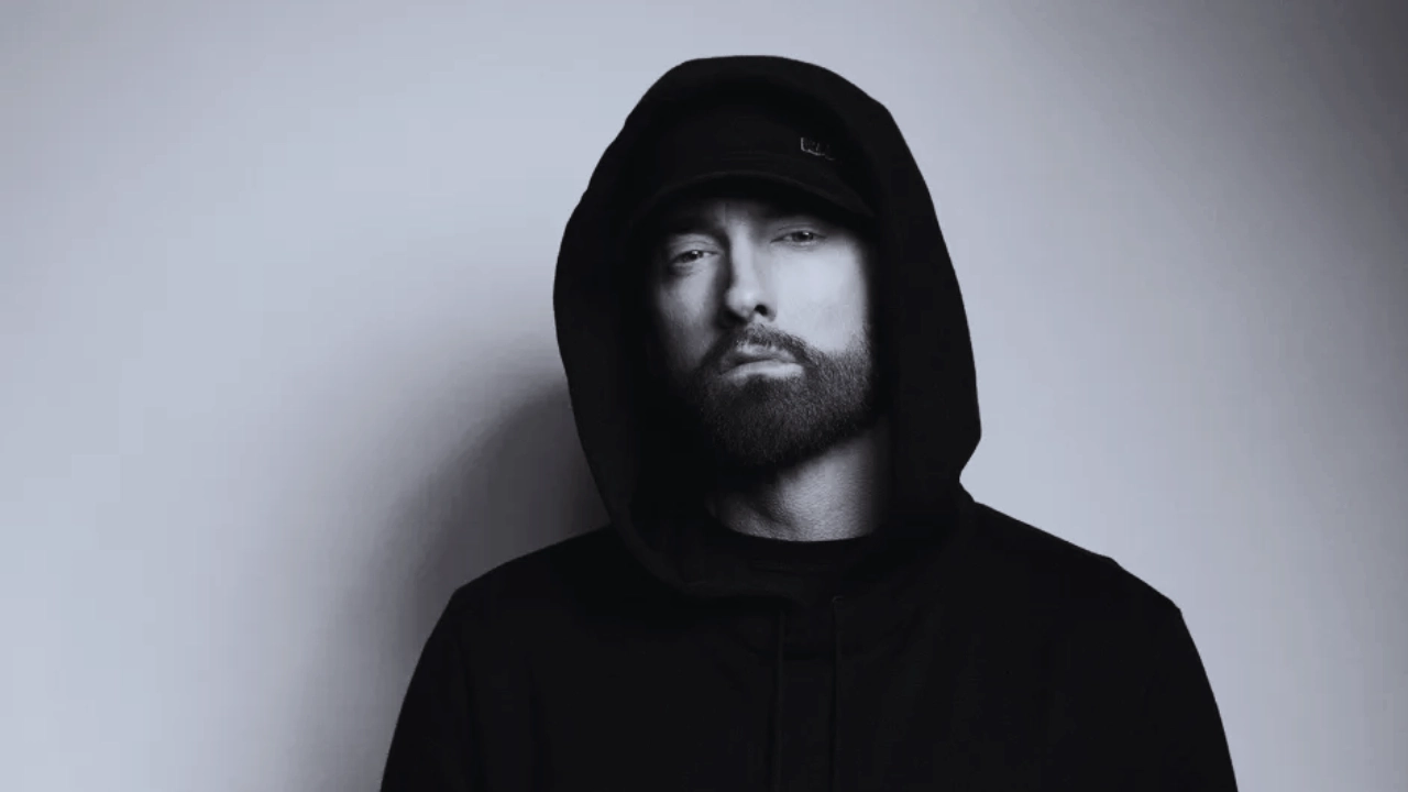 Eminem wearing a black hoodie and hat, staring directly at the camera with a serious expression.