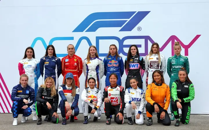 F1 Academy female drivers in team uniforms standing in front of F1 Academy banner.