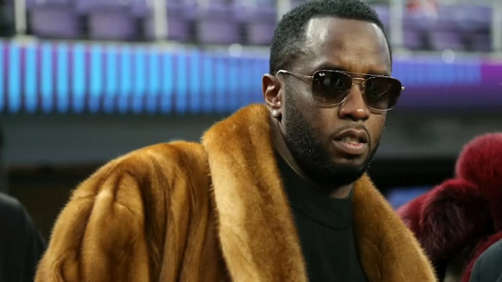 Sean Combs wearing a fur coat and sunglasses at an indoor event, looking serious.