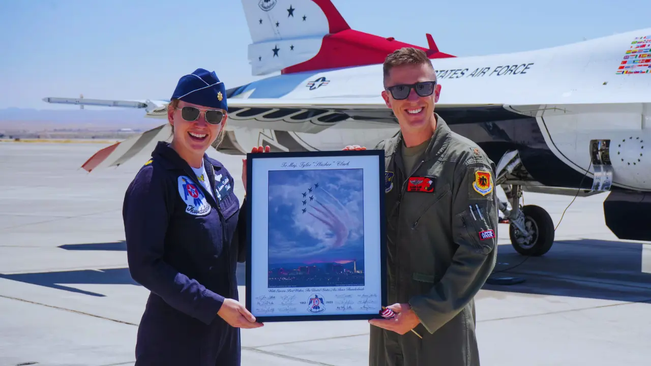 Major Lauren Schlichting and fellow Air Force pilot smile while holding a framed Thunderbird flight photo in front of a jet.