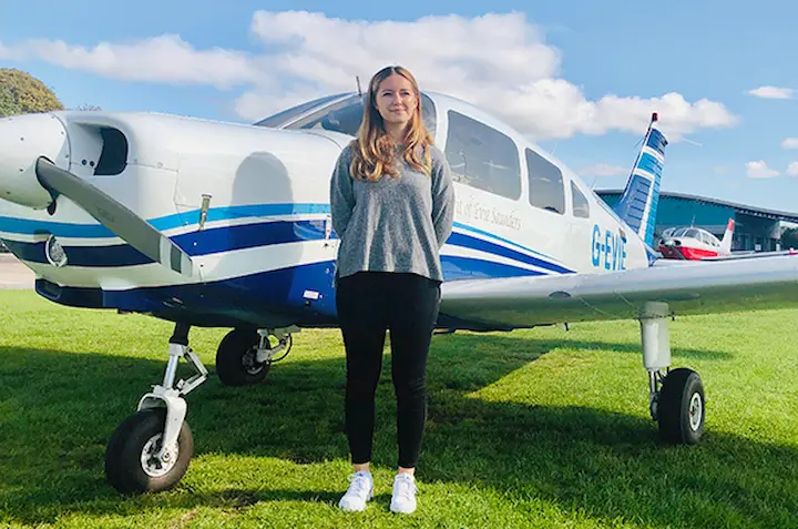 Lily Ayres standing next to a blue and white single-engine airplane on a sunny day.