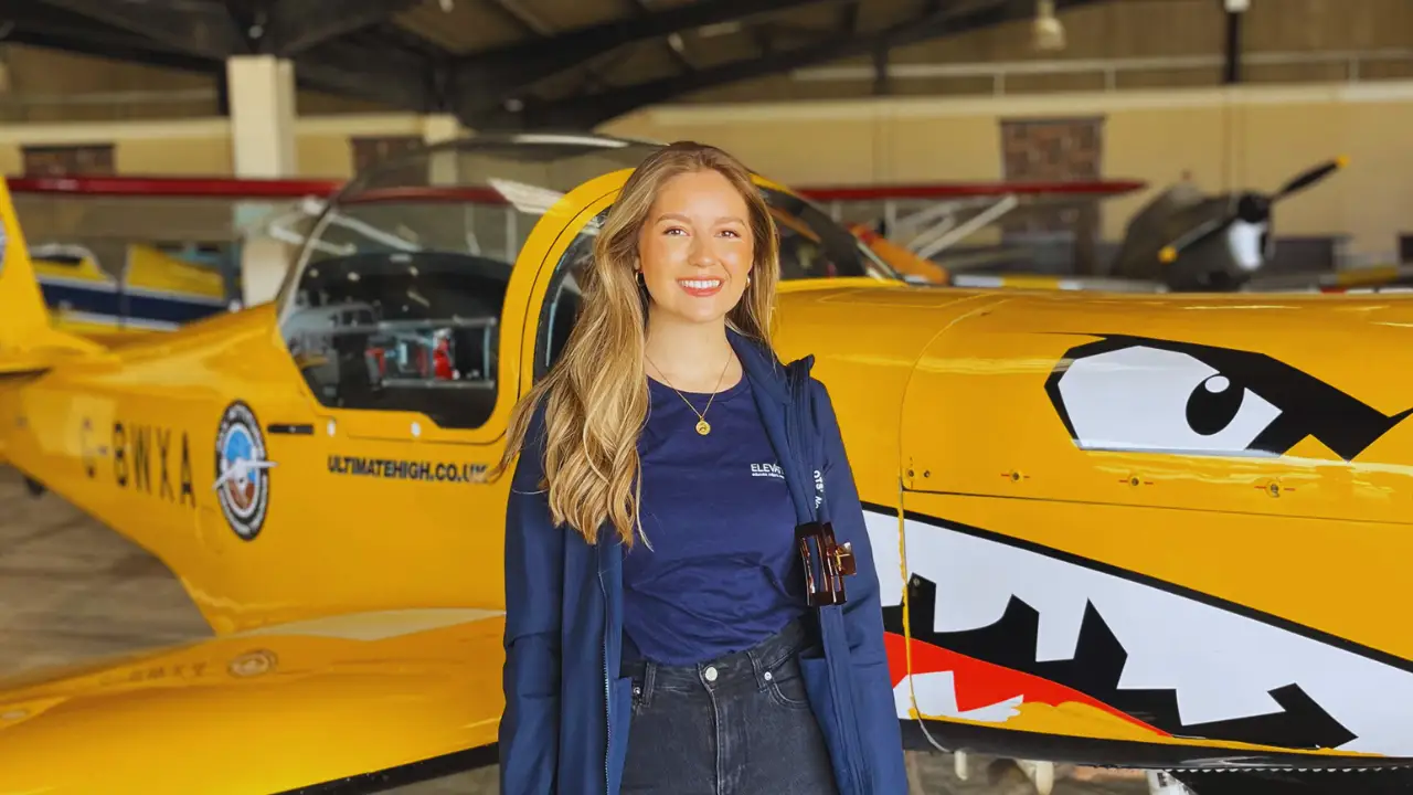 Lily Ayres smiling in front of a bright yellow stunt plane with painted shark teeth in an aircraft hangar.