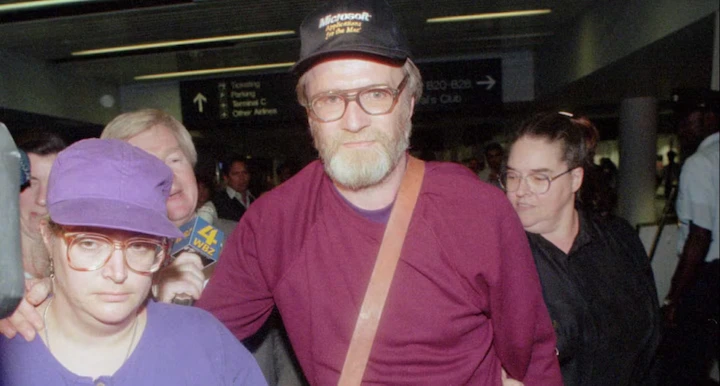 Suspect James Lewis wearing a cap and glasses at an airport, escorted by police.