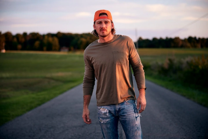 Morgan Wallen standing on a country road wearing a backward orange cap and casual clothes at sunset.