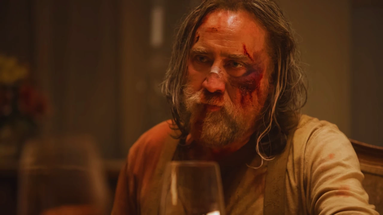 Nicolas Cage with a bruised face portraying Robin Feld in Pig, sitting at a table in a tense emotional moment.