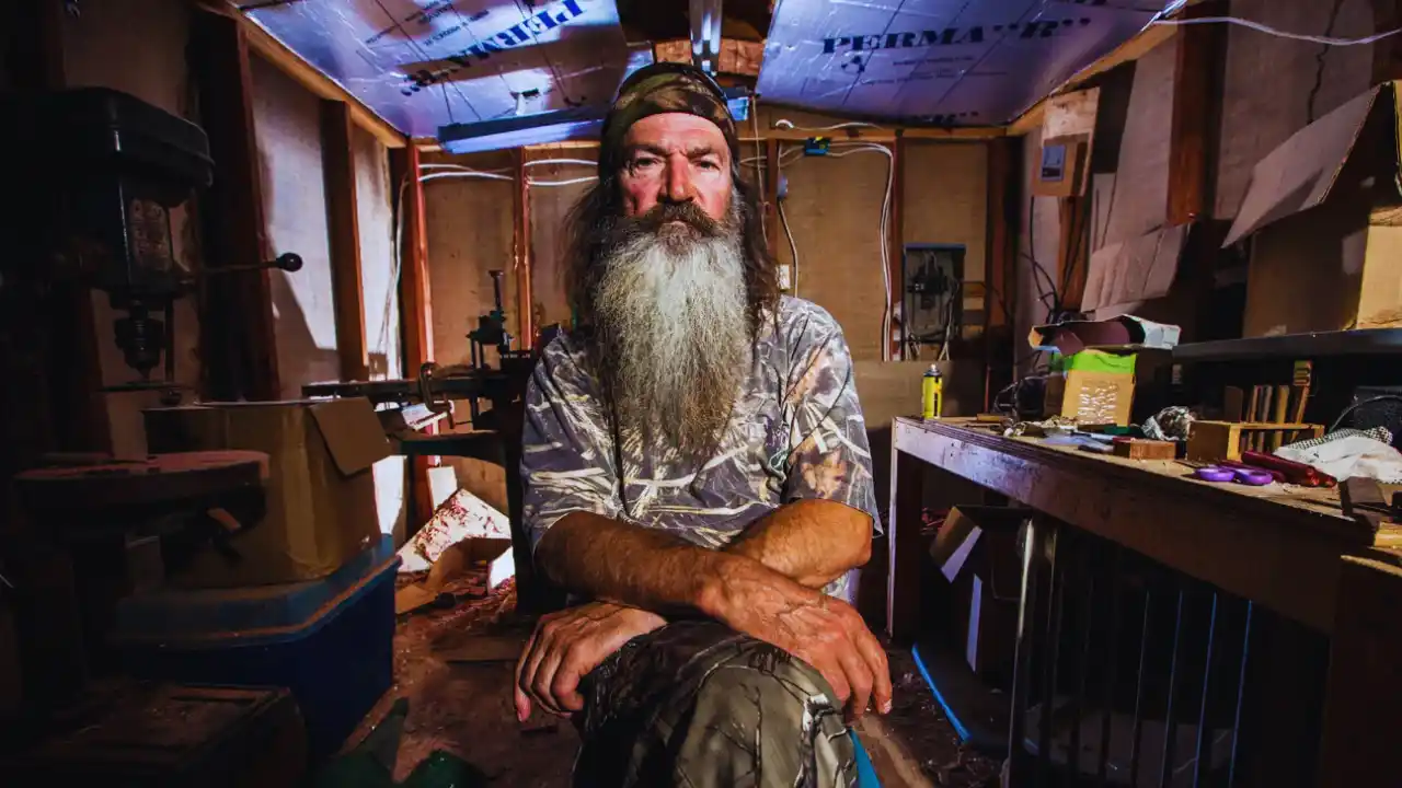 Phil Robertson sits in a rustic workshop, wearing camouflage clothing and a serious expression, surrounded by tools and wooden workbenches.