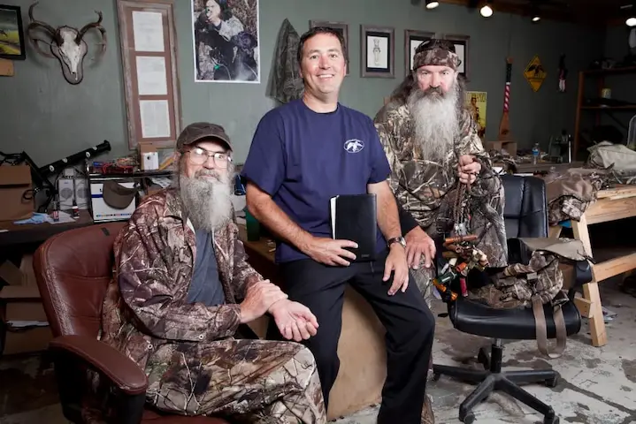 Phil Robertson poses with family members in a cozy office filled with hunting gear, symbolizing strong family bonds and traditions.