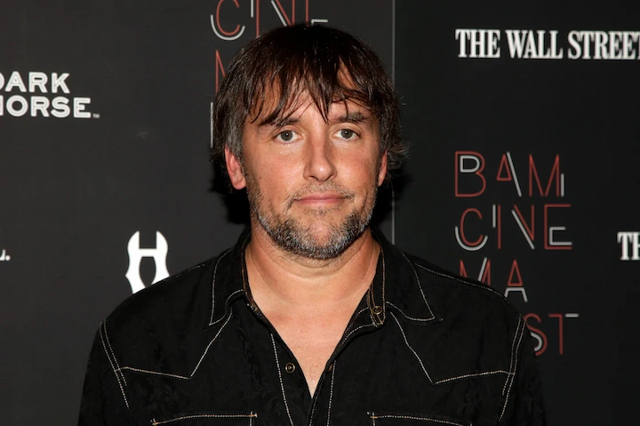 Richard Linklater attending a film premiere in a black shirt, standing in front of a sponsor backdrop.