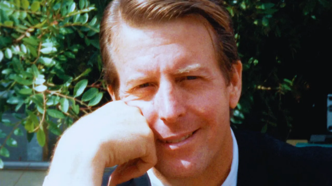 Richard Post smiling with his hand resting on his cheek, captured outdoors with green foliage in the background