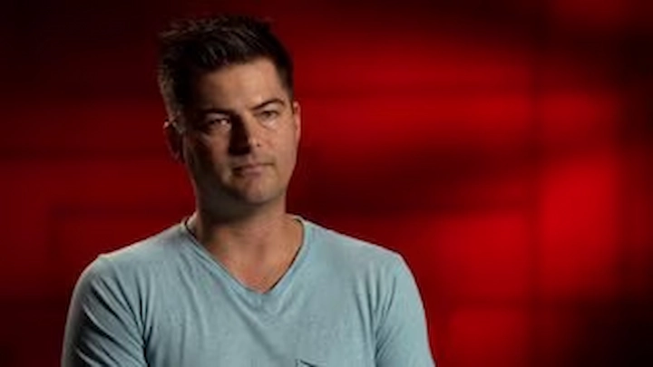 Richard Post’s adult son appearing in a somber interview, wearing a light blue shirt against a red background.