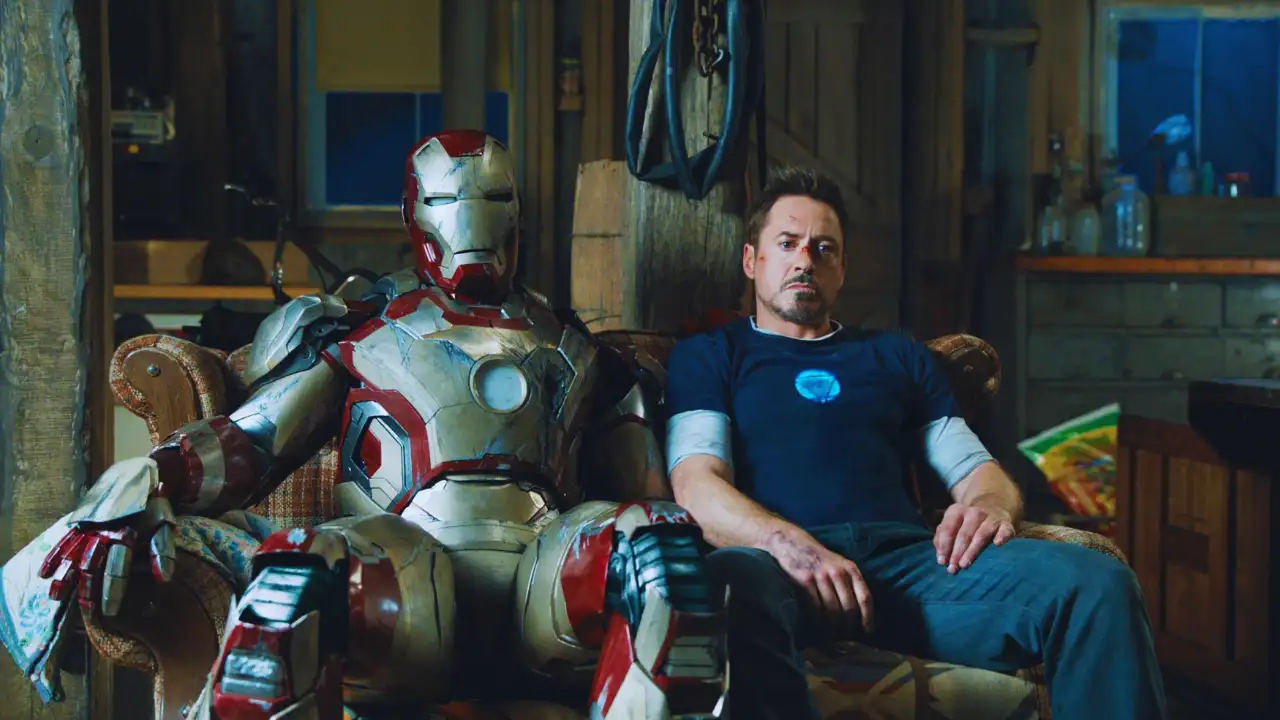 Robert Downey Jr. as Tony Stark sitting next to his Iron Man suit in a rustic cabin scene from Iron Man 3.