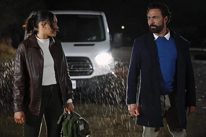A man and woman stand in the rain facing each other with a van and headlights behind them.