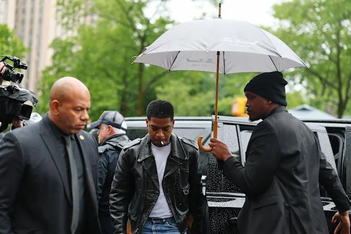 Kid Cudi walks toward court under an umbrella held by a security aide, wearing a black leather jacket and escorted by bodyguards.