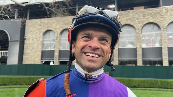Umberto Rispoli smiling in colorful jockey gear at the racetrack during a professional horse racing event.