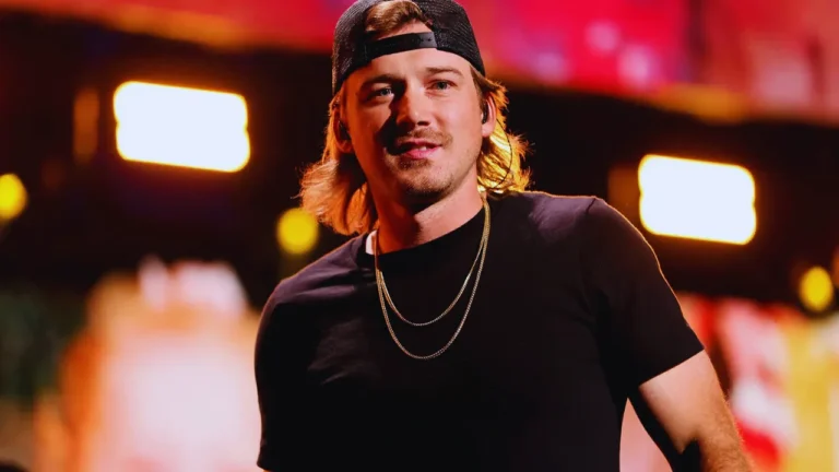 Morgan Wallen performing on stage in a black t-shirt and gold chains under dramatic lighting.