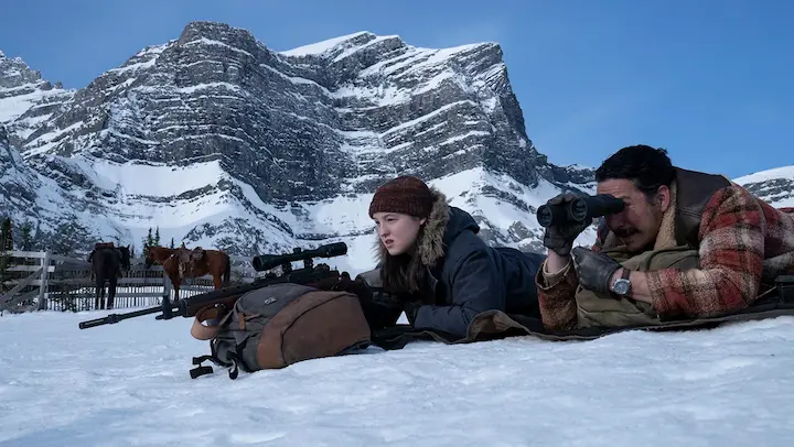 Ellie and Joel lying in the snow with rifles during a tense moment in The Last of Us Season 2, set against a backdrop of rugged mountains.