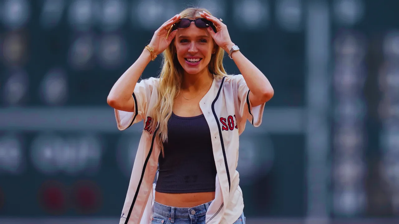 Alex Cooper smiling, wearing Boston Red Sox jersey, at a sports stadium event.