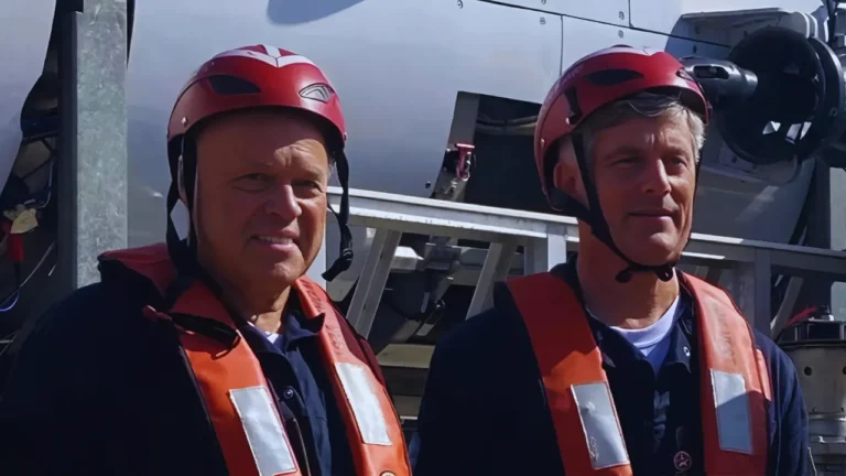 Alfred Hagen and Tym Catterson wearing red helmets and orange life vests standing beside the Titan submersible.