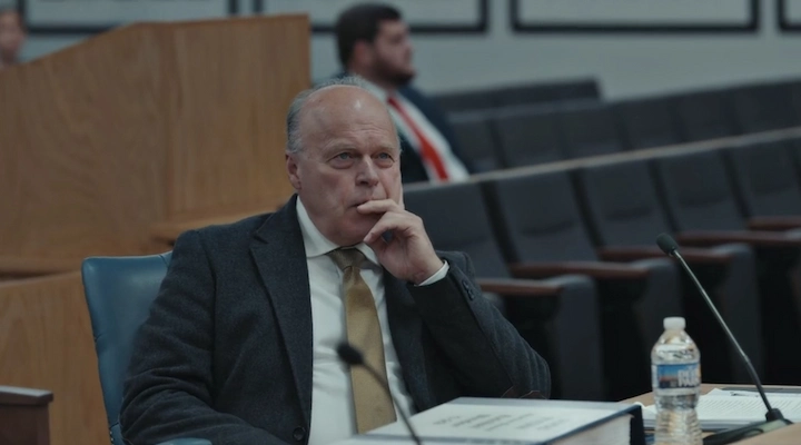 Tym Catterson testifying at a hearing, seated behind a microphone with a binder and water bottle, wearing a suit and gold tie.