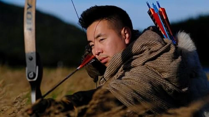 Contestant aiming bow and arrow in Alone Season 12 while lying in dry African grass.