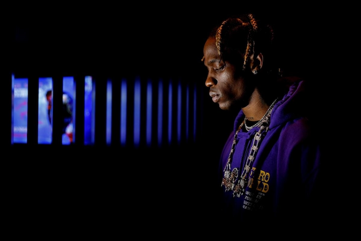 Travis Scott in purple hoodie stands in low light reflecting on Astroworld Festival tragedy.