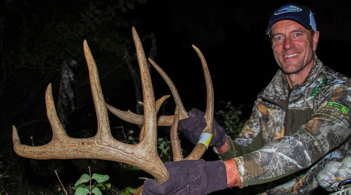 Bill Winke holding large deer antlers while wearing camouflage hunting gear and smiling at night