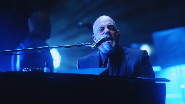 Close-up of Billy Joel singing into a microphone while playing piano amid soft blue stage lighting.