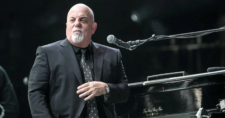 Mid-shot of Billy Joel in dark suit at grand piano hand on keys expressive pose under stage lighting.