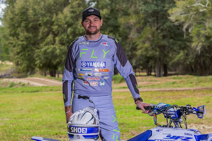 Chad Wienen in gray Yamaha FLY racing suit standing next to his ATV, outdoors on a dirt track.
