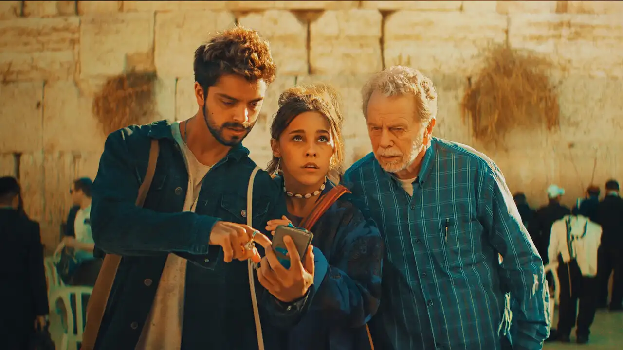Three characters from Cheers to Life examine a phone in front of an ancient stone wall.