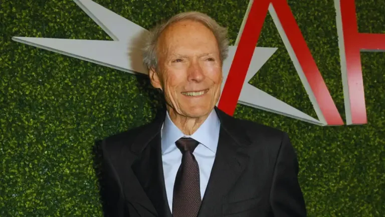 Clint Eastwood smiling at an AFI event, wearing a black suit and tie, with green backdrop behind him.