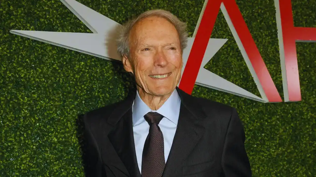 Clint Eastwood smiling at an AFI event, wearing a black suit and tie, with green backdrop behind him.