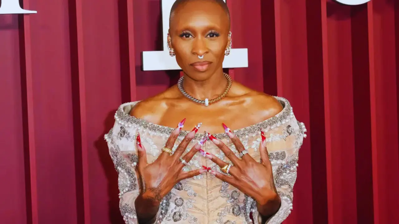 Cynthia Erivo in an off-the-shoulder beaded corset top and flowing peach skirt with red accent nail art.