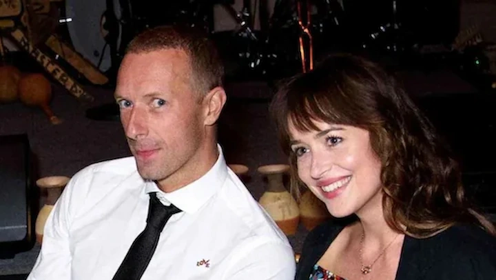 Dakota Johnson and Chris Martin seated together, smiling at an indoor event.