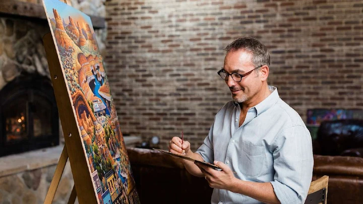 Eric Dowdle painting a colorful folk art scene indoors, capturing his creative process in the studio