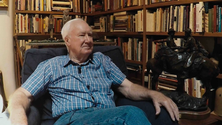 Forrest Fenn sitting in a chair surrounded by books and artifacts, reflecting on his adventurous life.